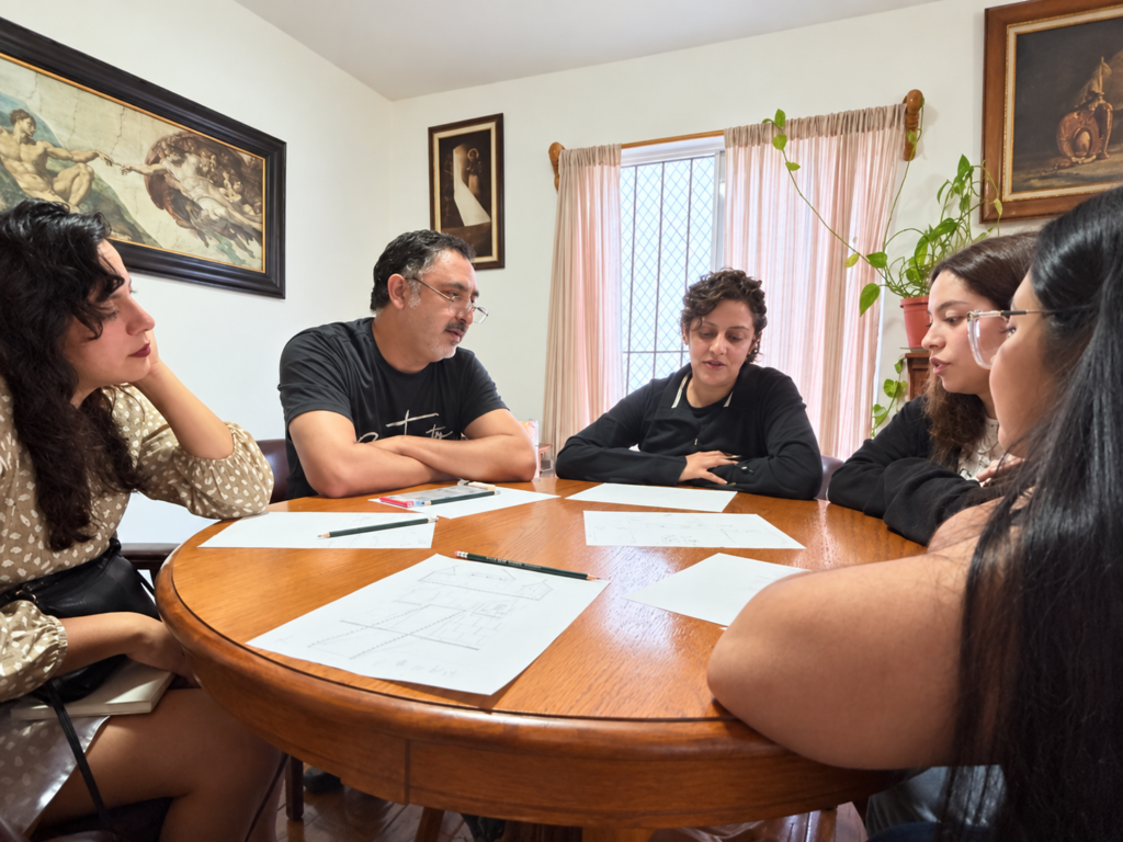 Cinco personas reunidas alrededor de una mesa de madera, compartiendo una actividad con hojas y lápices en Contacto.