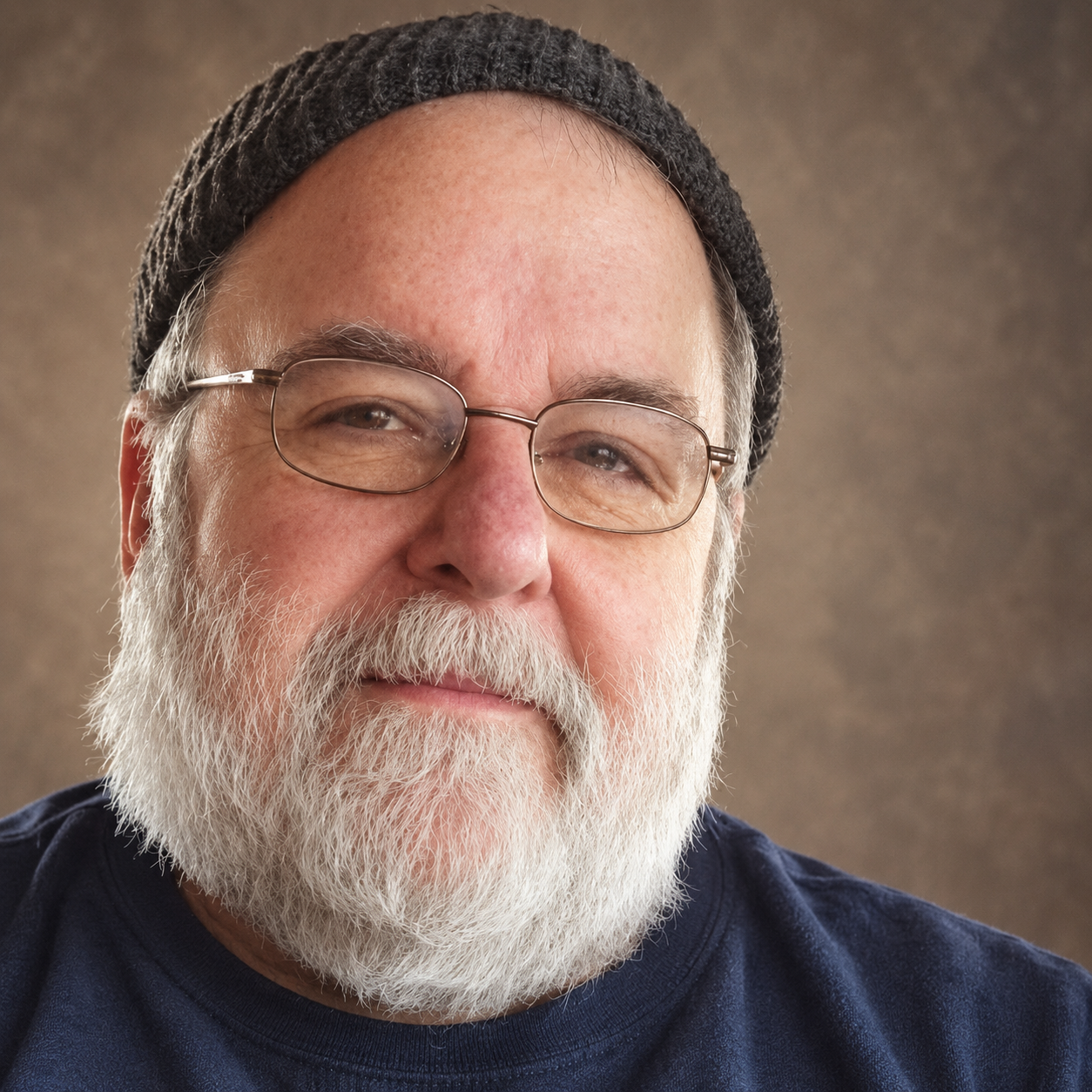 Retrato de Jon Jensen con lentes, barba blanca y gorro oscuro, sobre un fondo cálido de estudio.