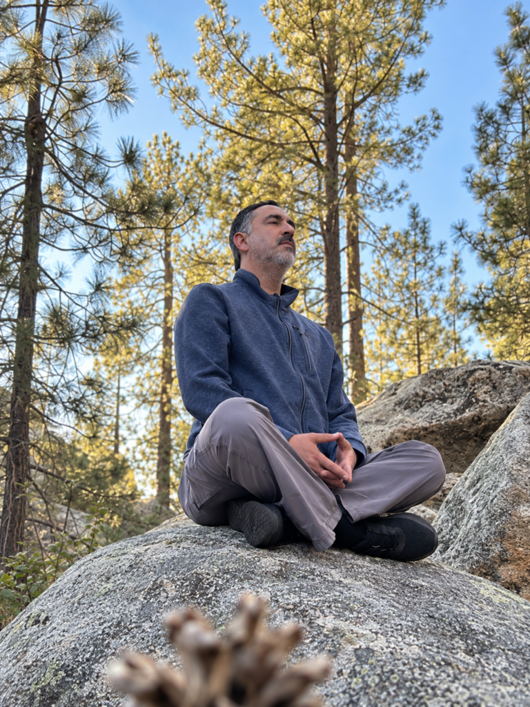 Hombre sentado con las piernas cruzadas sobre una roca en un bosque de pinos.