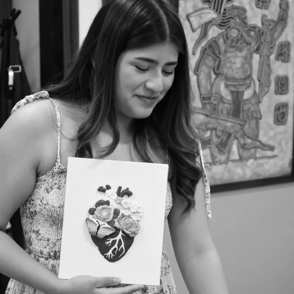 Participante sostiene un lienzo con un corazón anatómico en relieve decorado con flores y una mariposa, en blanco y negro.