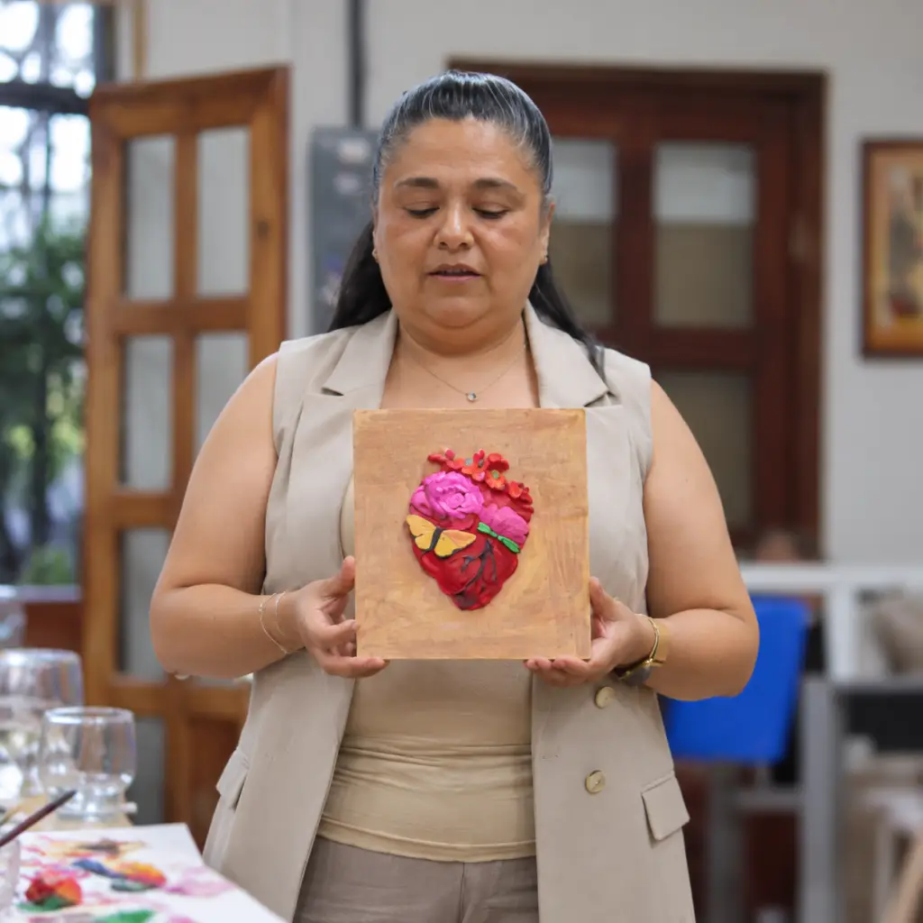 Mujer sostiene un cuadro de fondo dorado con un corazón rojo en relieve, flores rosas y una mariposa amarilla.